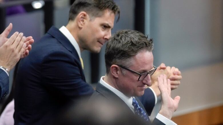 Two men in suits stand, one waving and one applauding.