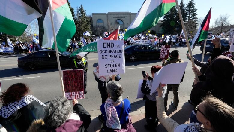 People protest on different parts of the street waving Palestine and Israel flags. Multiple signs are held, including a main one that says 