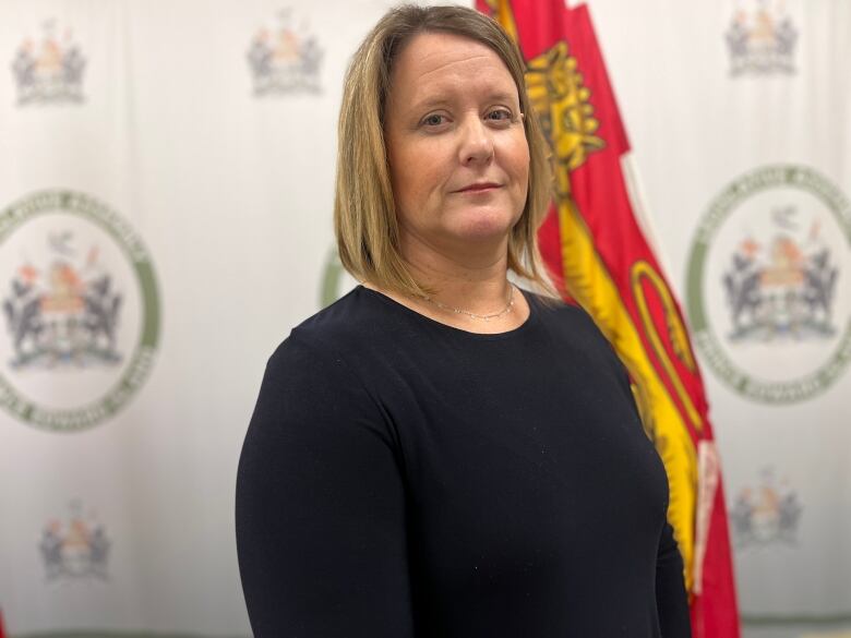 Jenn Redmond stands against a backdrop of flags. She is wearing a dark blue top and looks directly in to the camera.