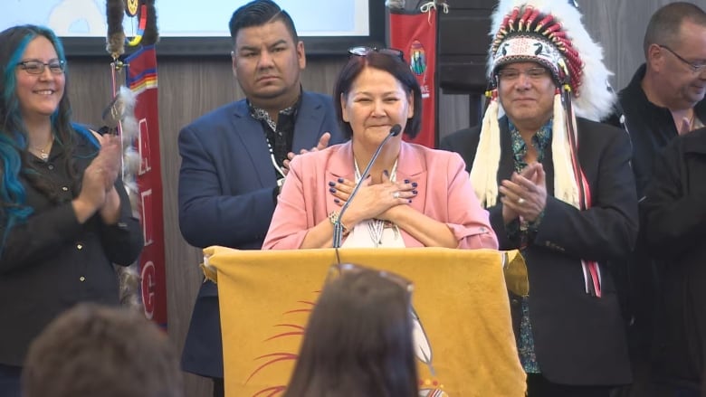 A woman stands at a podium, holding her hands to her heart, as people applaud.