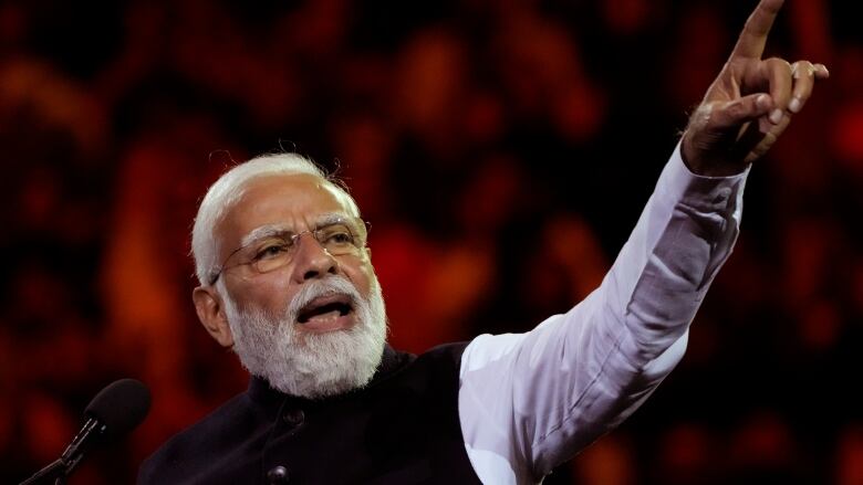 An Indian man with white hair and bear points a finger up in the air