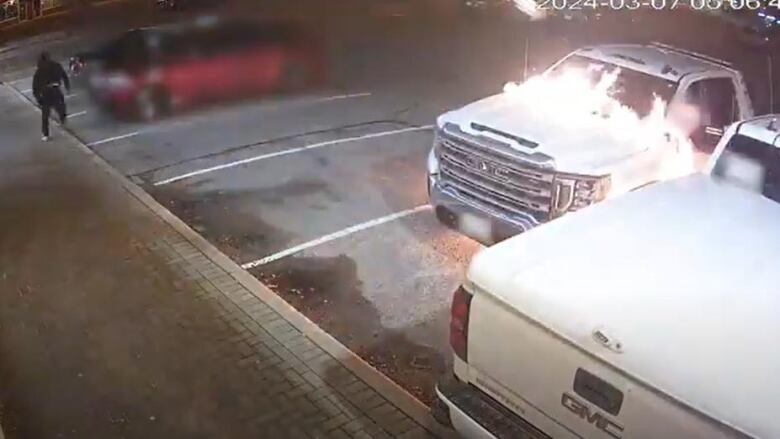 Surveillance footage showing a white tow truck in a plaza set on fire, and a suspect in dark-coloured clothing running away from the car. 