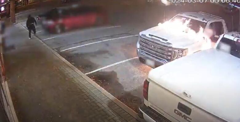 Surveillance footage showing a white tow truck in a plaza set on fire, and a suspect in dark-coloured clothing running away from the car. 