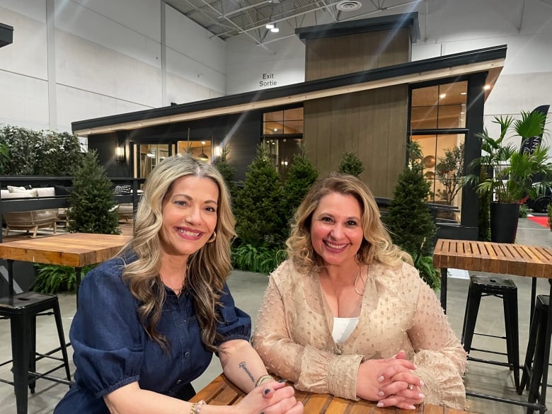 Two middle-age white, blond women are shown from the waist up, seated at a table in front a model home, in a convention centre.