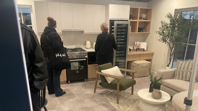 Three people, facing away from the camera, examine a kitchen and living room in a small residential unit