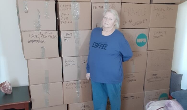 A woman wearing a blue shirt and pants stands in front a large stack of cardboard boxes in an apartment building.