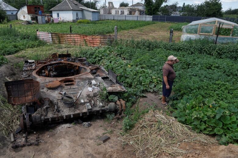 A man in a white cap stands to the right of a destroyed tank. They are in a garden. 