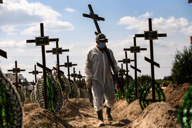 A man wearing white protective gear walks through a graveyard holding a cross. There are rows of crosses and wreaths behind him marking graves.