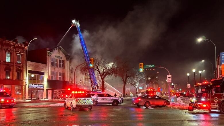 Emergency vehicles fill street, as a ladder truck directs water down onto a burning building.