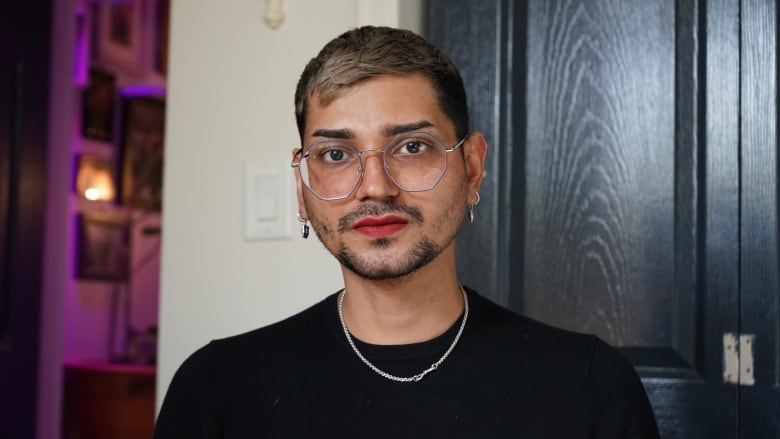 Portrait of a man with big round glasses, silver jewelry, lipstick, with soft lighting in the background. He has a serious expression.