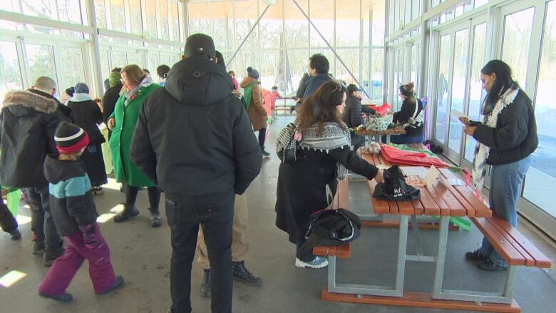 A group of people inside a glass-walled shelter includes people at tables with kite supplies on them.