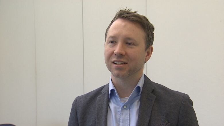 A man in a grey jacket and blue shirt speaks to the camera