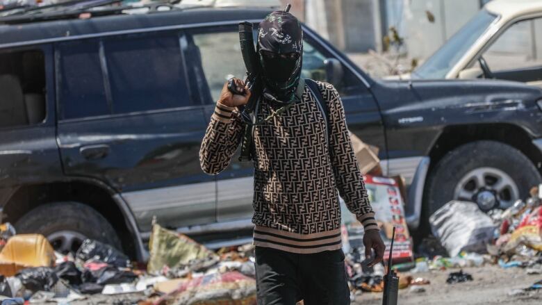 An armed gang member wearing a face mask walks down a street with a gun over his shoulder.