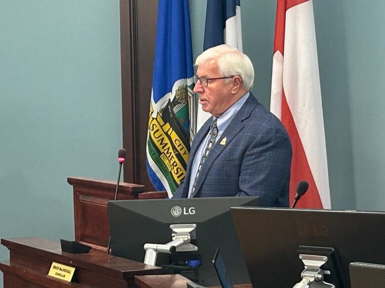 A man in a blue suit jacket stands at a podium with flags behind him.