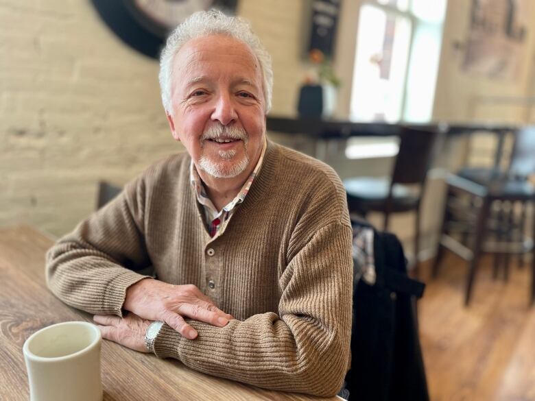 A man in a brown sweater and grey hair sits at a cafe table.