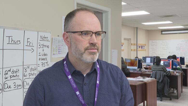 A man wearing a button up shirt and glasses stands with a classroom behind him.