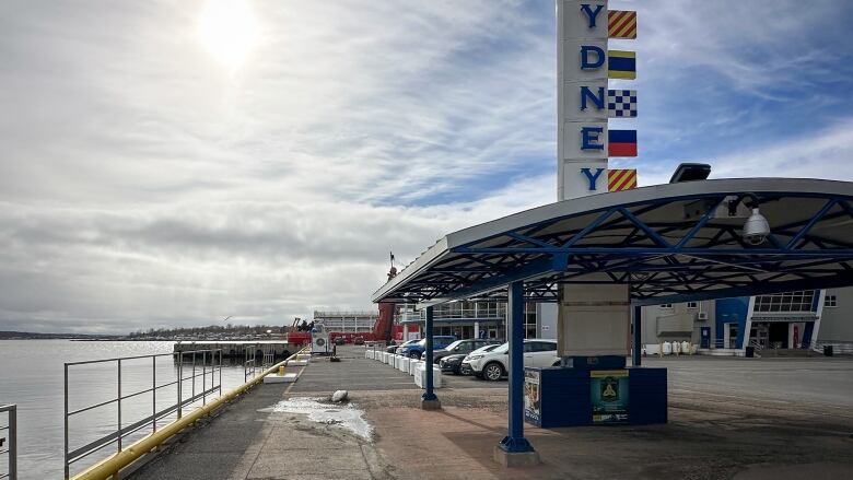 A concrete dock on the water with cars parked off to the side, a covered area so people can get out of the sun or rain, and a large vertical sign saying SYDNEY. 