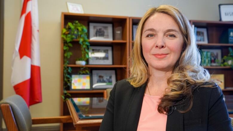 A blonde woman in a dark suit stands in an office with a Canadian flag in the background.