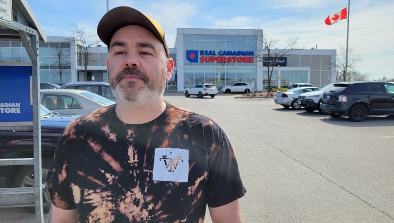 A man in a baseball hat and tie-dye shirt stands in a parking lot, outside a supermarket.