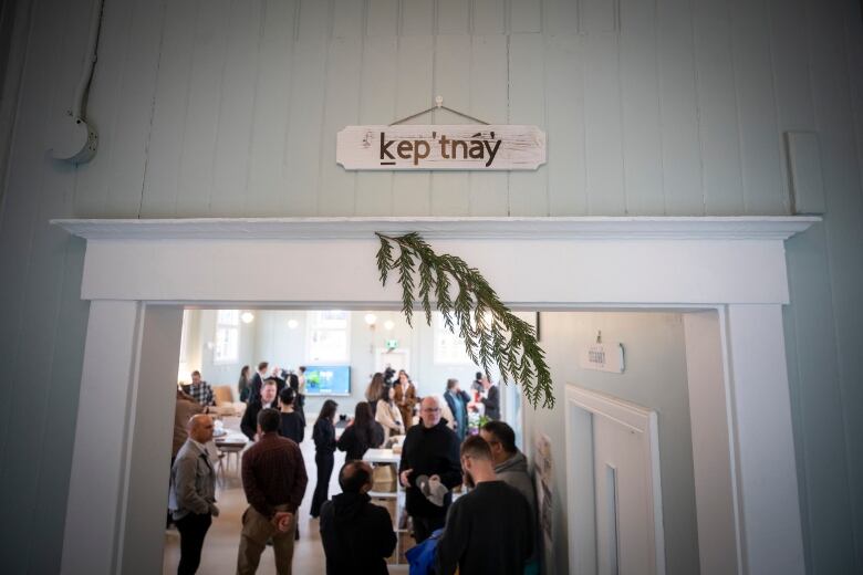 A sign in Squamish that says kep'tnay with a cedar bough hangs over a doorway. The room beyond is filled with people.