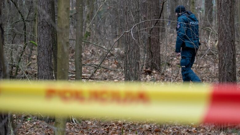 A man in a blue police uniform walks in a wooded area, with red and yellow police tape out of focus in the foreground.