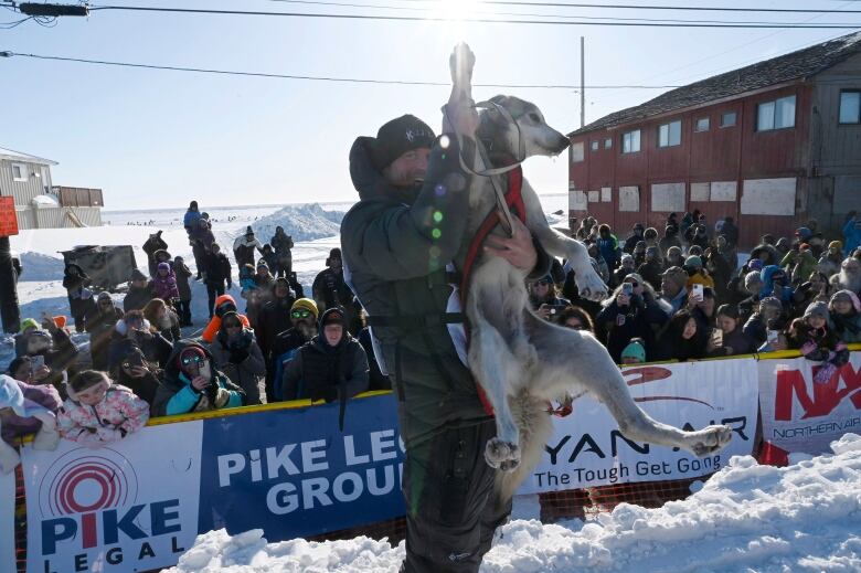 A man holds up a dog on a snowy street as a crowd of people looks on from behind a barrier.