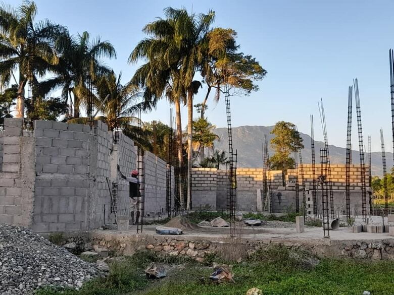 A man works on a small building of cement blocks. Palm trees are in the background.