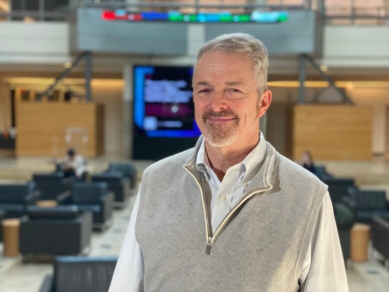 A man in a quarter-zip vest and white shirt stands in a university atrium. 