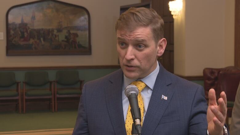 Man with blonde hair, wearing a blue suit taking to a microphone.