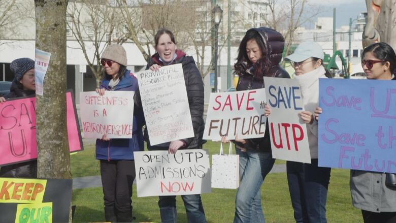 People are standing in a line wearing jackets and toques. They are holding colorful signs, some of which read 