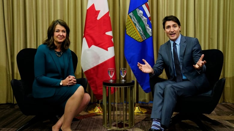 Man holds out both hands while talking, as woman sitting next to him smiles.