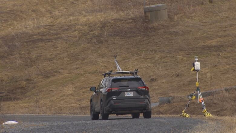 A black SUV with a metal antennae arm on the roof drives on a gravel road toward a concrete well. Beside the car is a large metal tripod with scientific equipment on top.