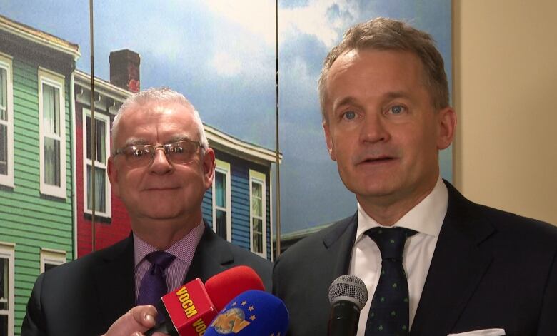 Two men stand in front of a city backdrop with microphones in front. 