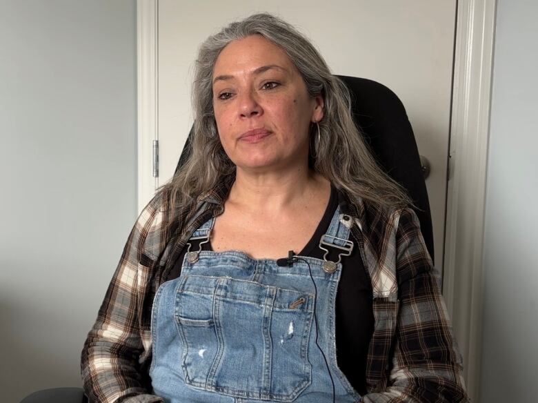 A person with long grey hair wearing overalls sits in front of a door.