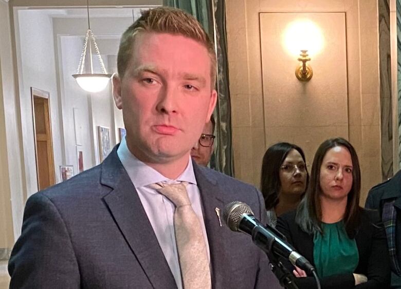 A man in a grey suit and a cream tie stands at a microphone while other people are behind him listening.