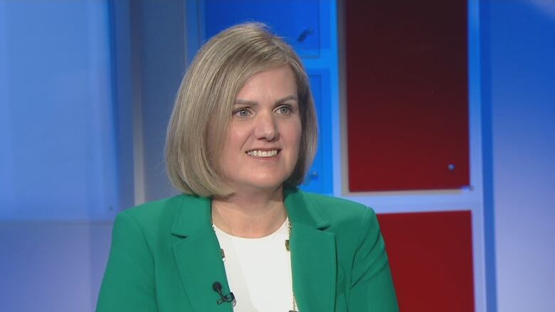 A woman in a green blazer and white shirt sits in a television studio.