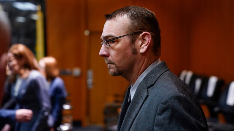 A man with a trimmed beard, wearing glasses and a grey suit, walks into a courtroom.