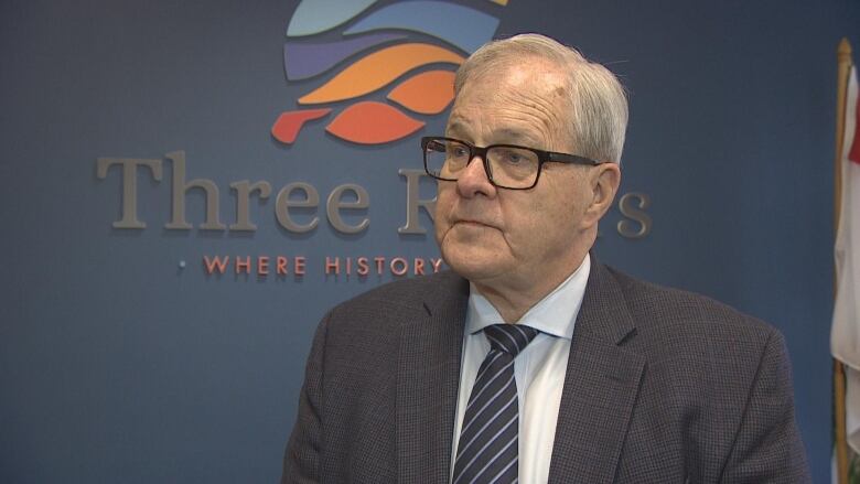 A man with glasses, grey hair, and wearing a suit stands in front a sign saying Three Rivers. 