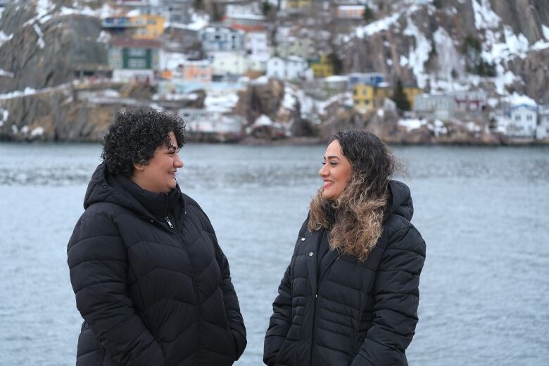 Two women face each other with water visible in the background.