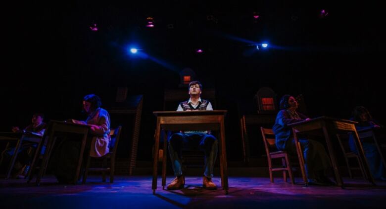 A performance stage with four actors sitting behind wooden desks. The male performer in the centre is looking into the distance. 