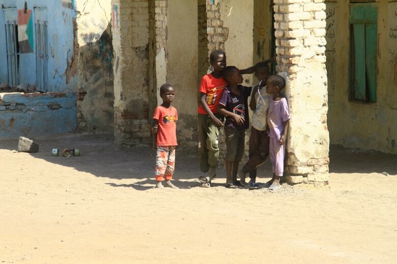 Four young boys stand outside a building in the sun next to a brick pillar. 