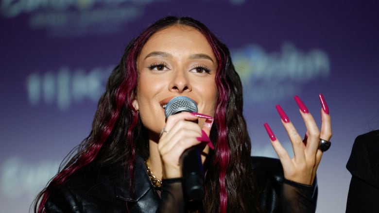 A woman with long, dark hair, smiles and holds a microphone. 