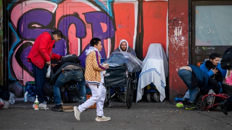 People line up on a sidewalk in front of a graffiti wall.