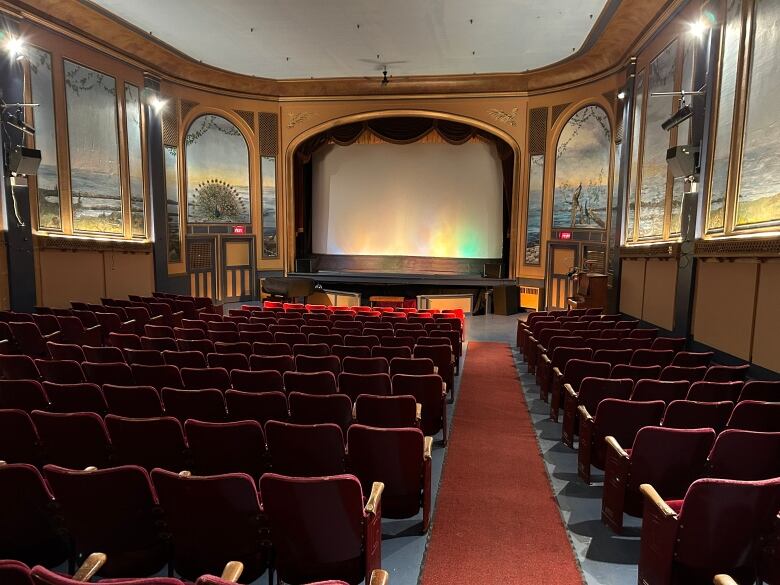 A white screen is lit up in an auditorium with rows of red chairs, a walkway with red carpet, and walks lined with painted murals of trees and wildlife. 