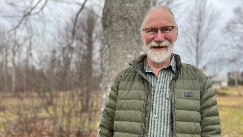 Man with grey beard standing outdoors in front of tree.