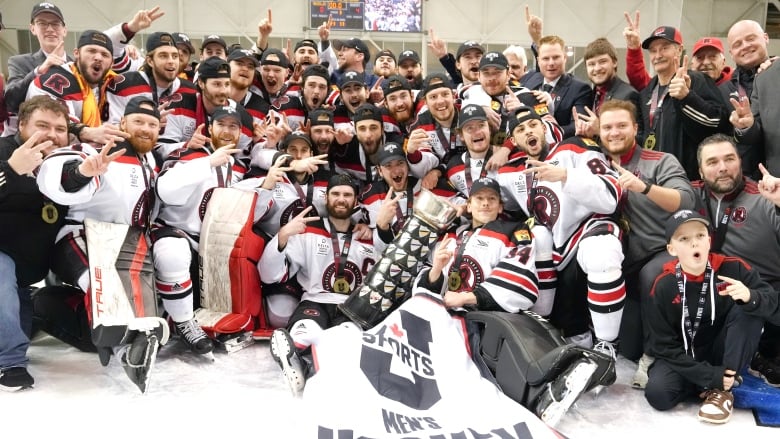 A male ice hockey team poses on the ice with a trophy and banner.