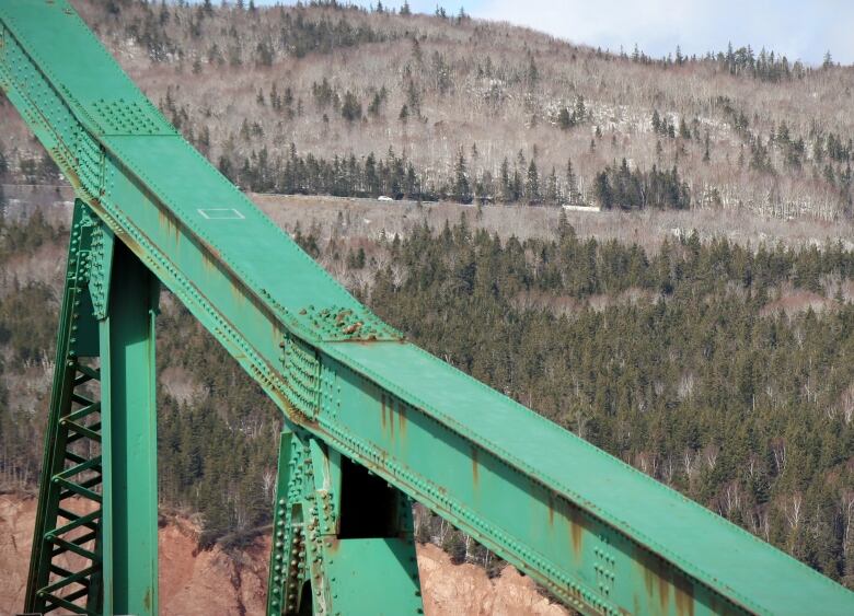 Green steel beams of a bridge in the foreground looking up at a hill covered in trees, with a silver tanker seen in the distance at the edge of a road,