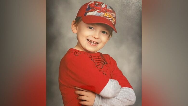 A boy wears a Spider-Man hat and a red t-shirt with a gray long sleeve shirt underneath. His arms are folded across his chest.