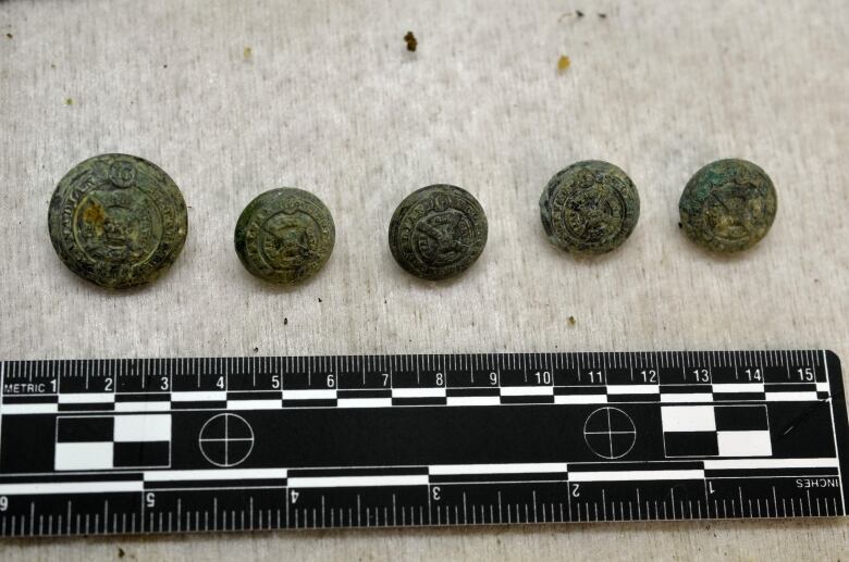 Five weathered buttons are shown against a ruler, indicating they are only a few inches wide. Some clearly show 