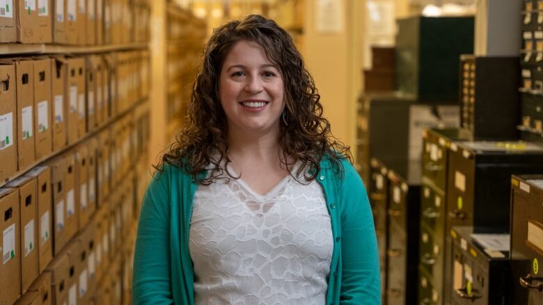 A woman in a cardigan and lace top smiles. She's standing in the aisles of filing cabinets.